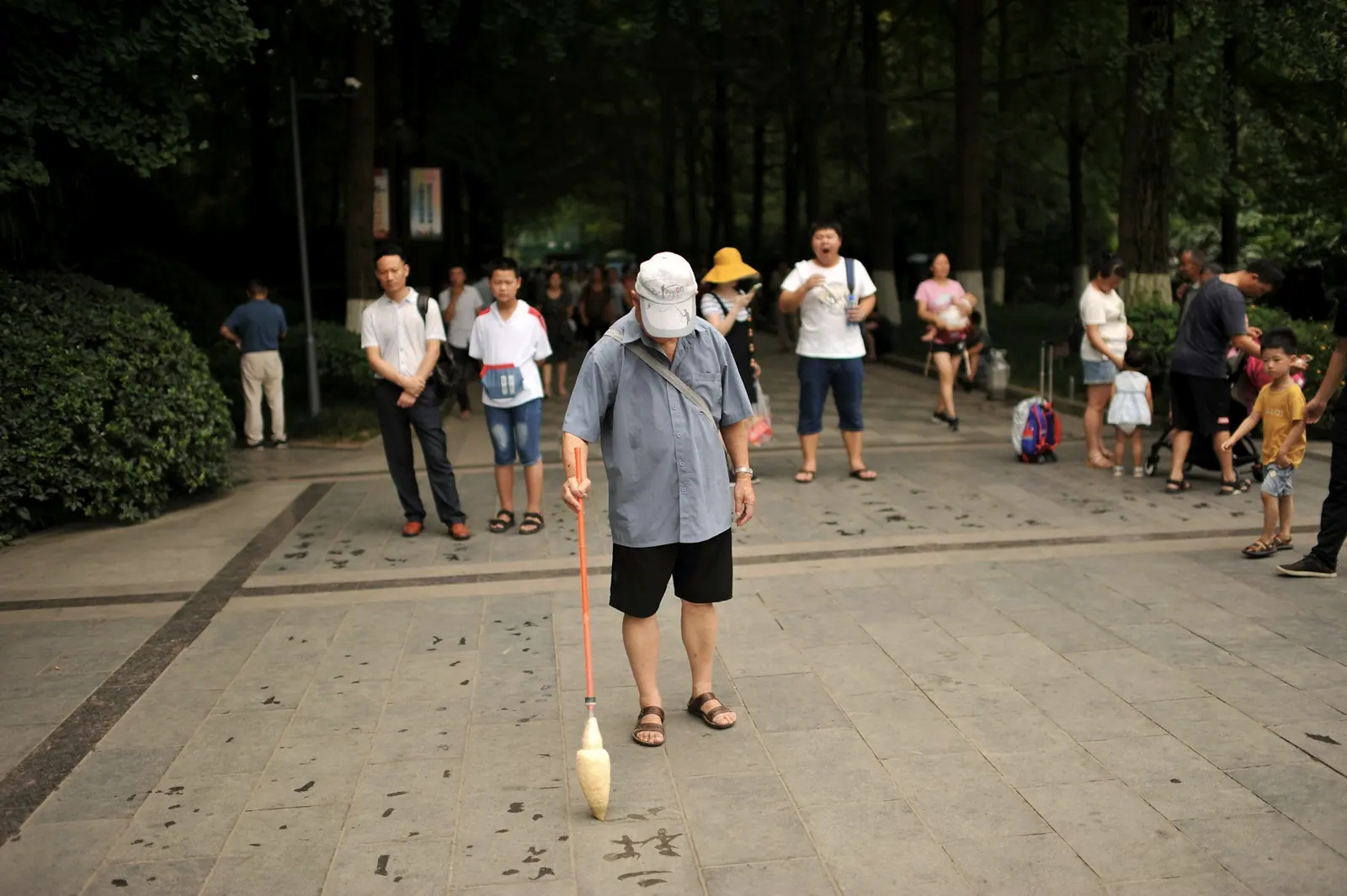 一位老人拿着长杆在广场地砖上写水字，旁边围着几个看的人